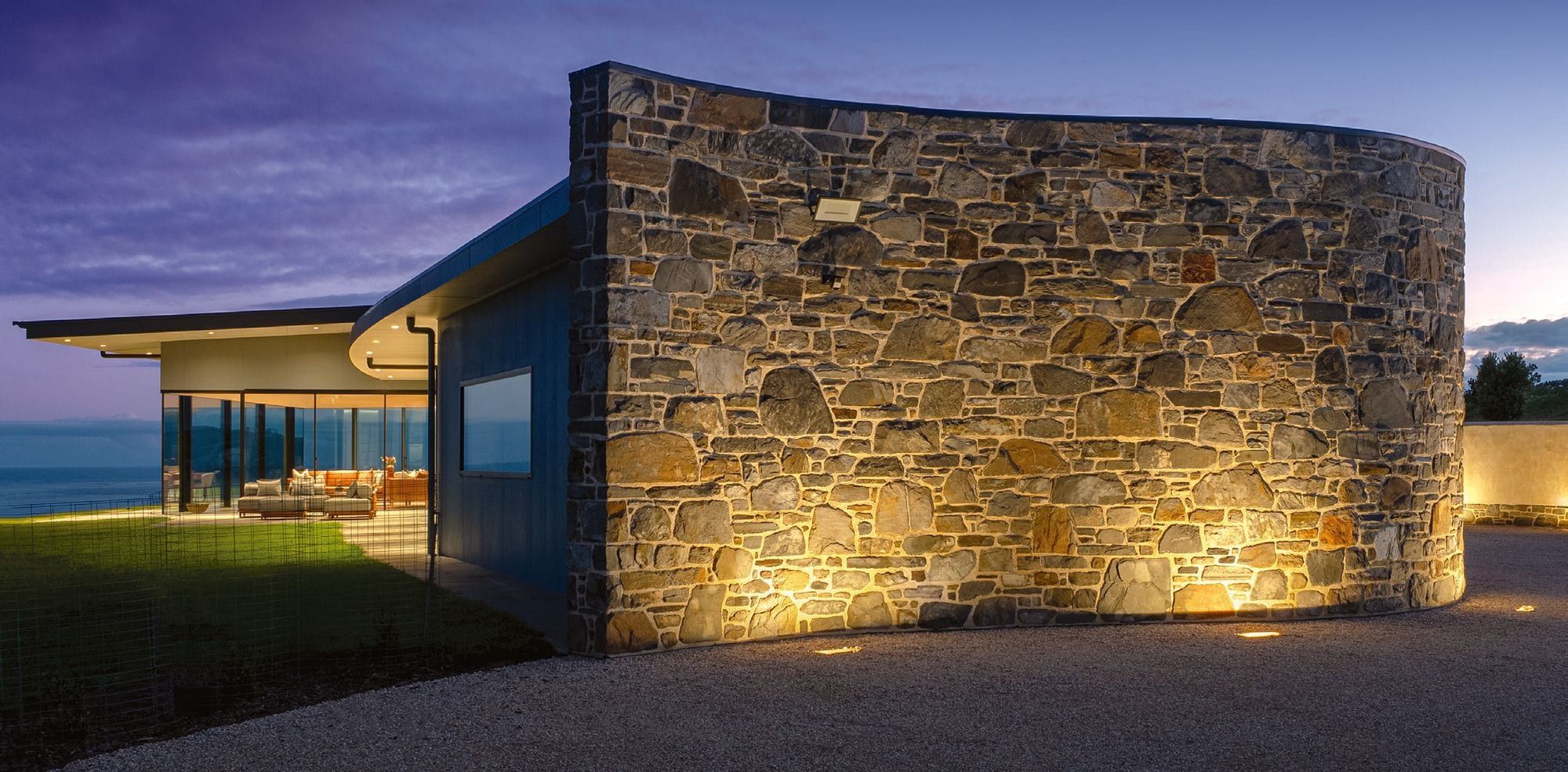 A curved stone wall lit at dusk beside a modern clifftop home with the ocean beyond